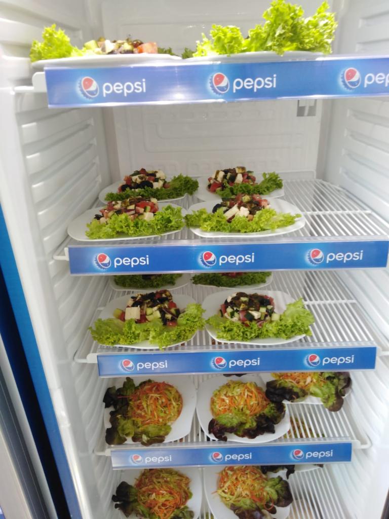 Salatki w lodówce z logo Pepsi na półkach, różne rodzaje sałatek ułożone na białych talerzach z zieloną sałatą, widoczne składniki: pomidory, ogórki, ser feta, marchewka.