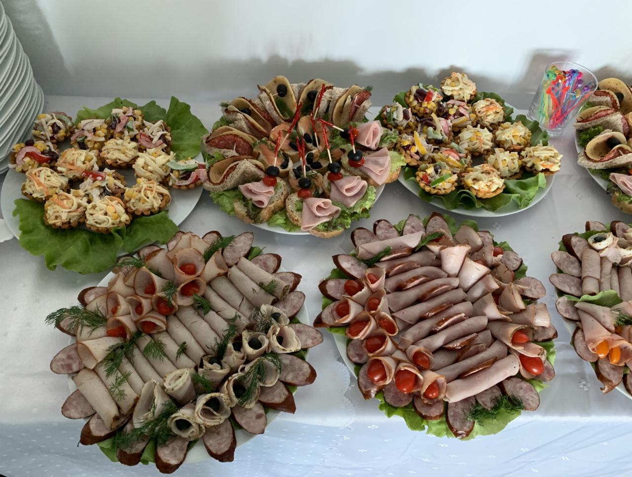 Dekoracyjne talerze z przekąskami na białym stole: tartaletki z sałatką, kanapki z szynką i oliwkami, roladki z wędliną i pomidorkami cherry, udekorowane zielonymi liśćmi sałaty i koperkiem.