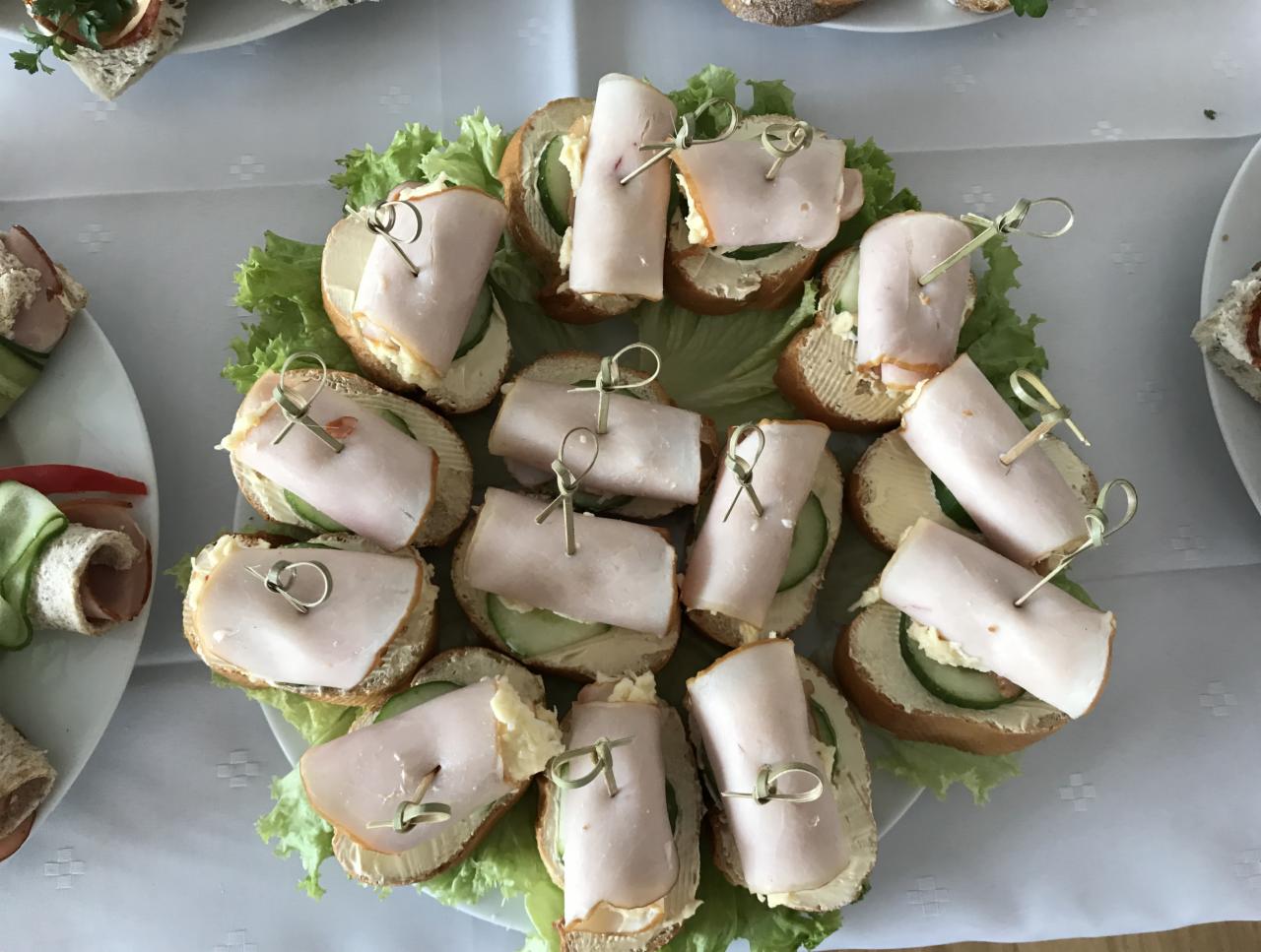 Talerz z małymi kanapkami na jasnym pieczywie z kremowym smarowidłem, ogórkiem i zwiniętym plasterkiem szynki, spięte ozdobnymi wykałaczkami, ułożone na liściach sałaty, widok z góry.