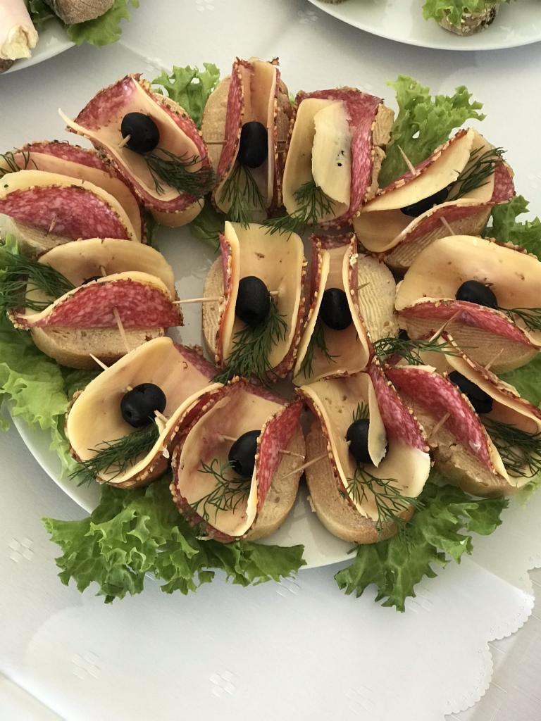 Dekoracyjnie ułożone na talerzu, okrągłe kanapeczki z salami, serem, czarną oliwką i koperkiem, otoczone liśćmi sałaty.