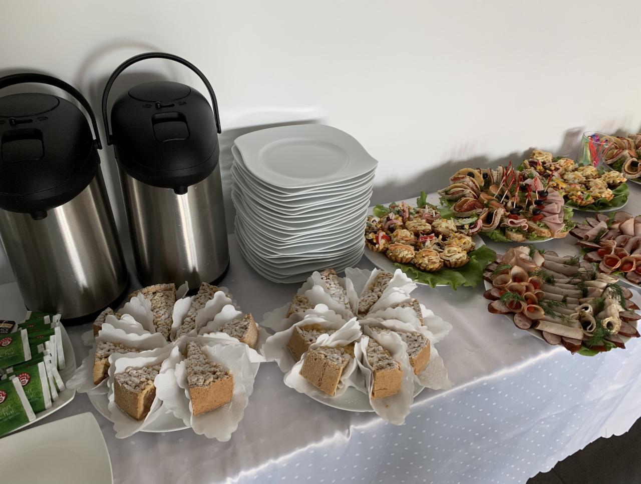 Stół z białym obrusem, na którym znajdują się dwie srebrne termosy, stos białych talerzy, oraz talerze z przekąskami: kanapeczki, tartaletki, wędliny i ciasto w kształcie trójkątów. Widoczne...