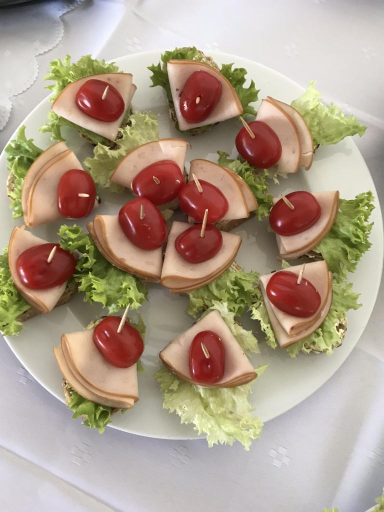 Talerz z apetycznymi kanapeczkami: ciemne pieczywo, świeża sałata, plaster wędliny i pomidorek koktajlowy, spięte wykałaczką, na białym obrusie.