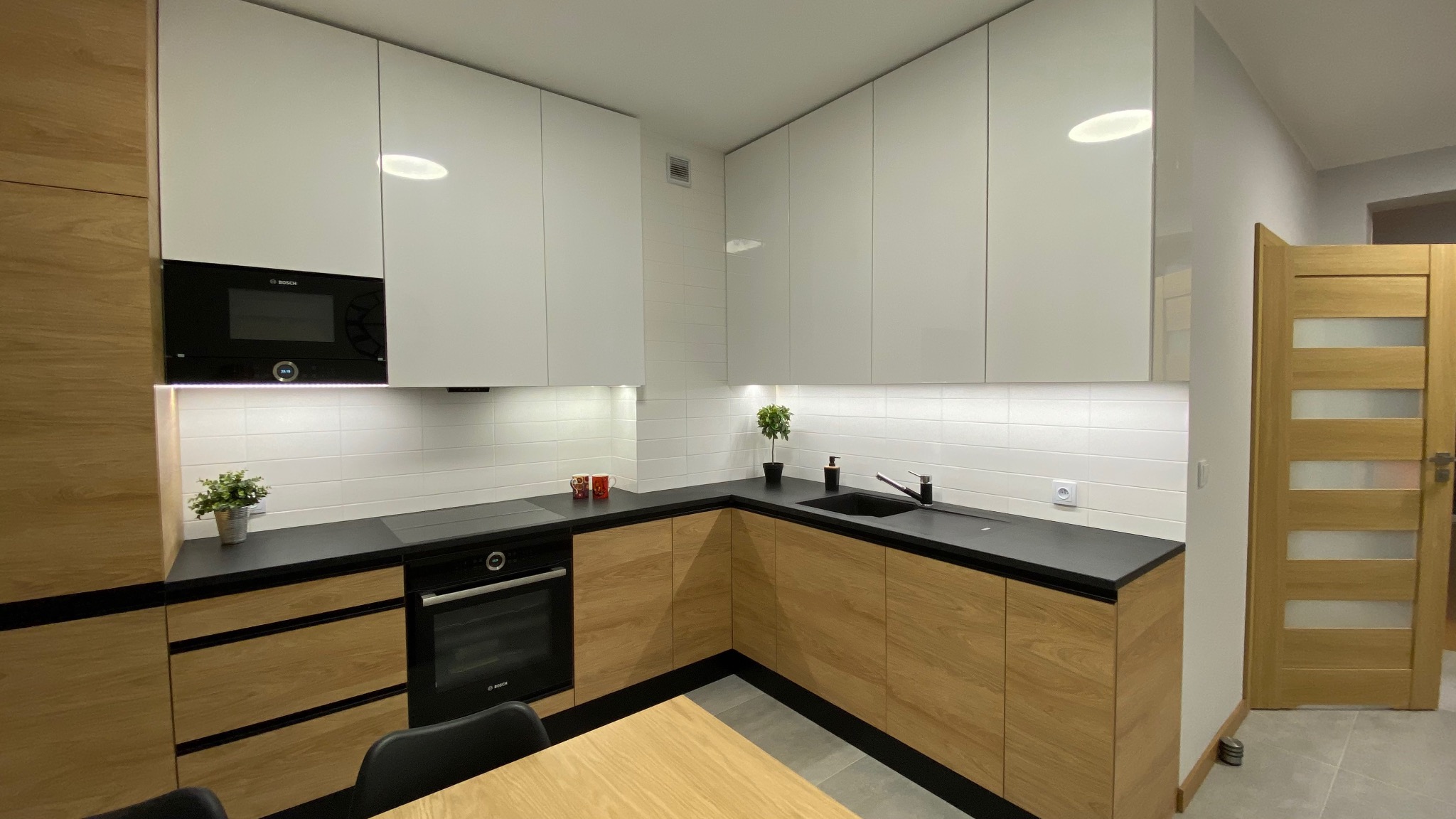 Nowoczesna kuchnia z białymi szafkami górnymi i drewnopodobnymi szafkami dolnymi, czarnym blatem, białymi kaflami na ścianie i wbudowanym piekarnikiem Bosch.