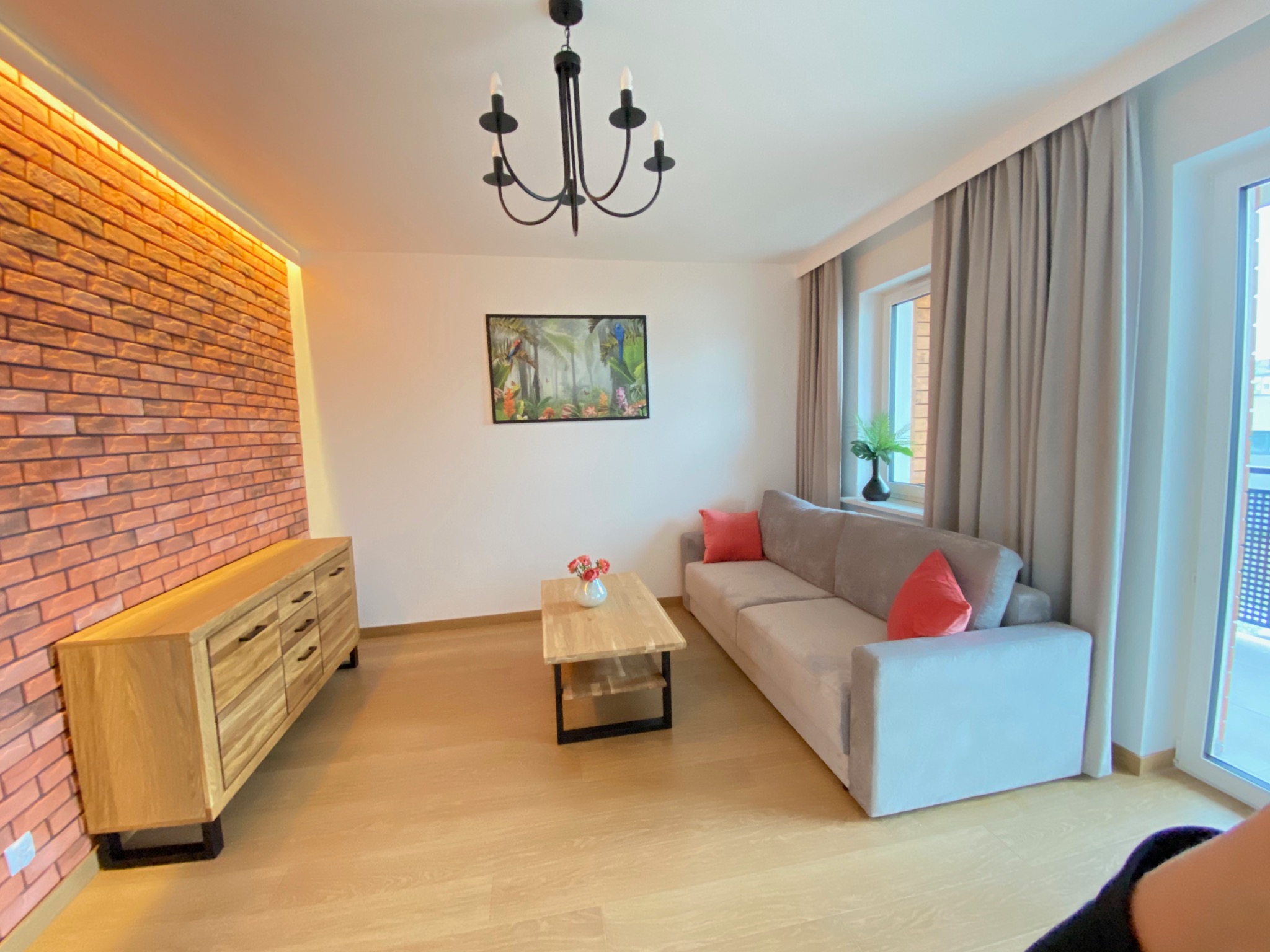 Nowoczesny salon z ceglaną ścianą z podświetleniem LED, drewnianą komodą, szarą sofą z pomarańczowymi poduszkami, drewnianym stolikiem kawowym i czarnym żyrandolem.