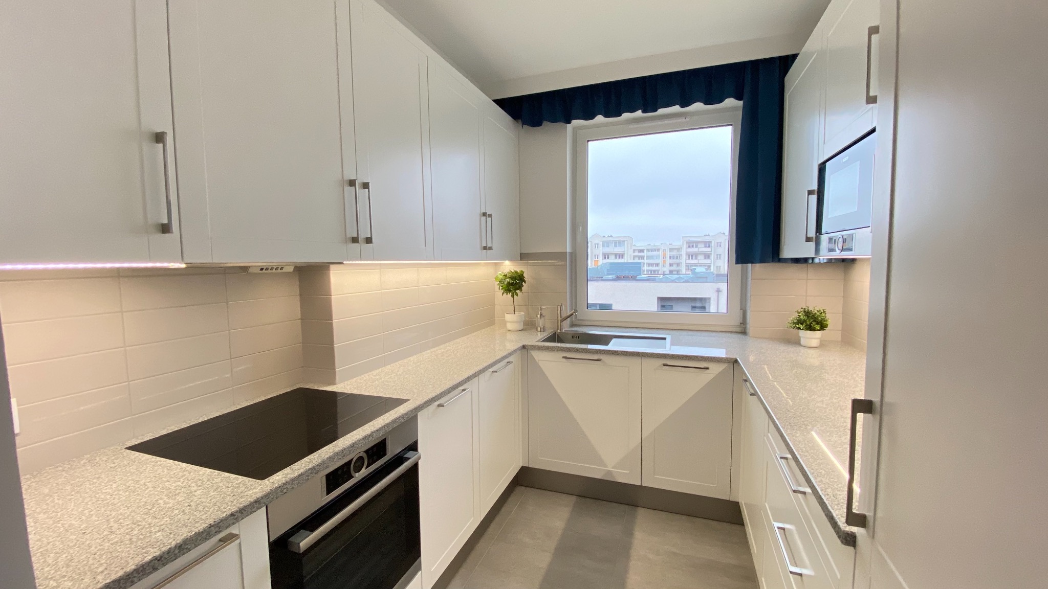 Jasna, nowoczesna kuchnia z białymi szafkami, szarym blatem i płytą indukcyjną. Nad blatem podświetlenie LED. Na parapecie doniczka z małym drzewkiem. Za oknem widok na miejski krajobraz Płocka.