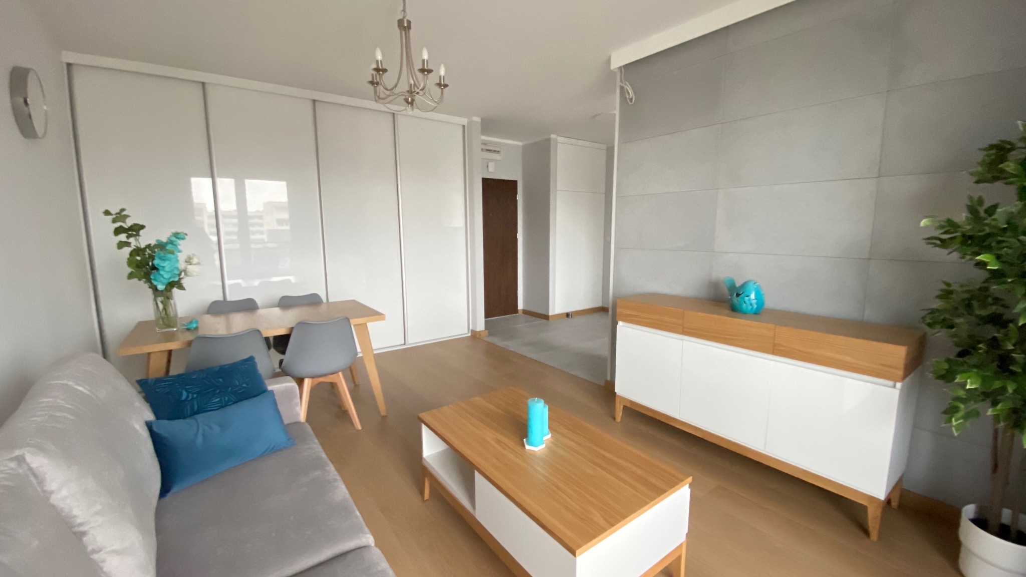 Jasne wnętrze salonu z szarym narożnikiem, drewnianym stolikiem kawowym z turkusowymi świecami, stołem jadalnym z szarymi krzesłami i białymi szafami przesuwnymi w tle, uzupełnione o dekoracje...