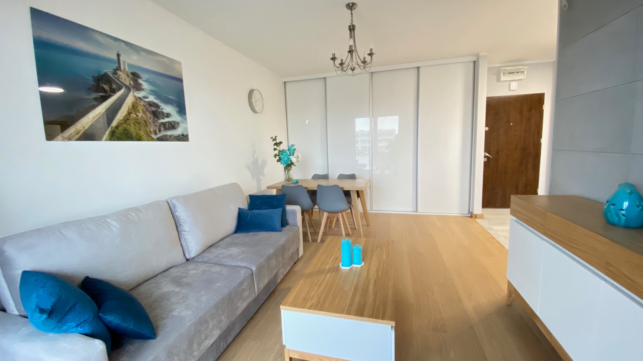 Jasne wnętrze mieszkania w Płocku: szara sofa z turkusowymi poduszkami, drewniany stół z szarymi krzesłami, obraz z motywem morskim na ścianie, drewniana podłoga i białe szafy przesuwne.