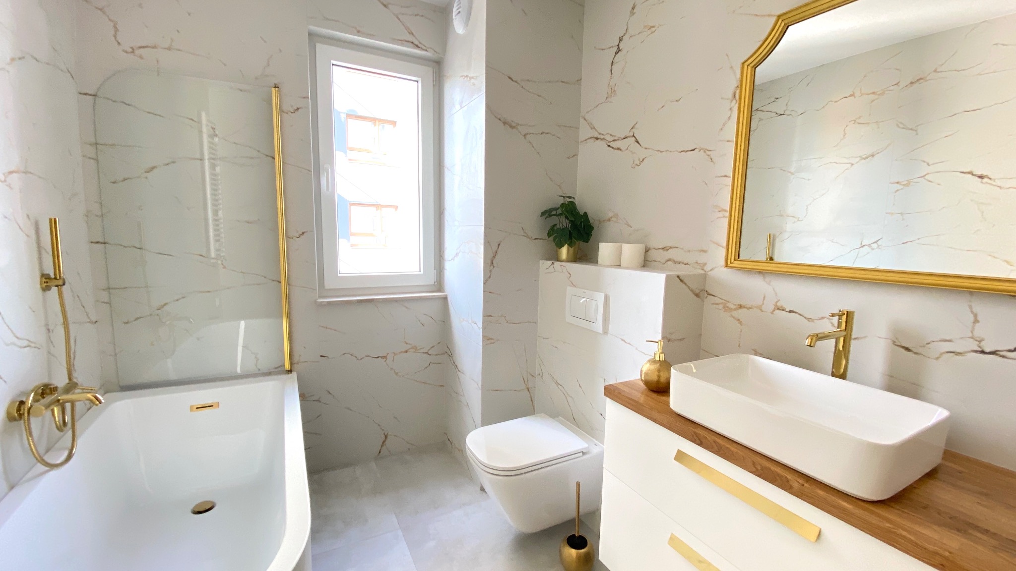 Elegancka łazienka z białymi marmurowymi płytkami, złotymi akcentami armatury i dodatków, jasnym drewnianym blatem pod umywalką oraz oknem z widokiem na budynek.