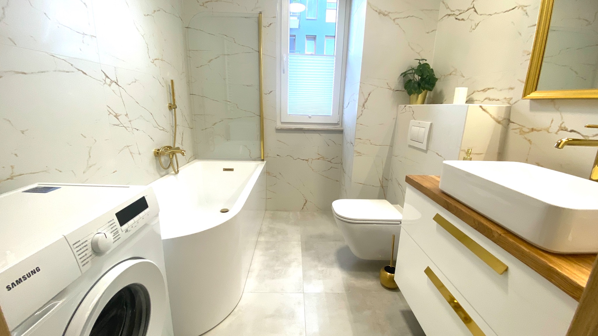 Biała łazienka z elementami w kolorze złota: wolnostojąca wanna, umywalka nablatowa, wisząca toaleta, pralka Samsung i płytki imitujące marmur.
