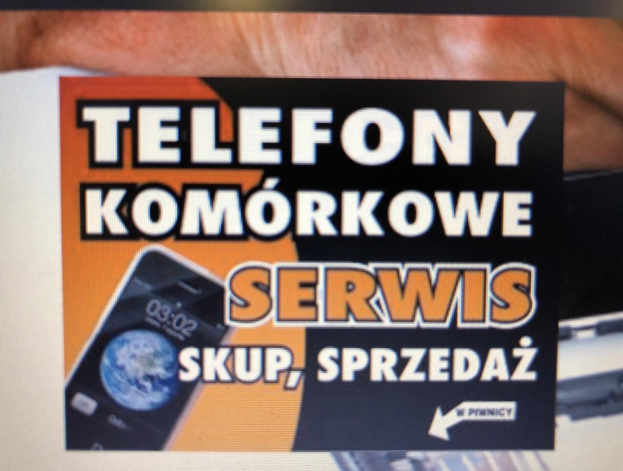 Billboard reklamowy z napisem 'Telefony komórkowe Serwis Skup, Sprzedaż' i grafiką smartfona z wyświetlonym globusem, wskazujący strzałką 'W piwnicy'.