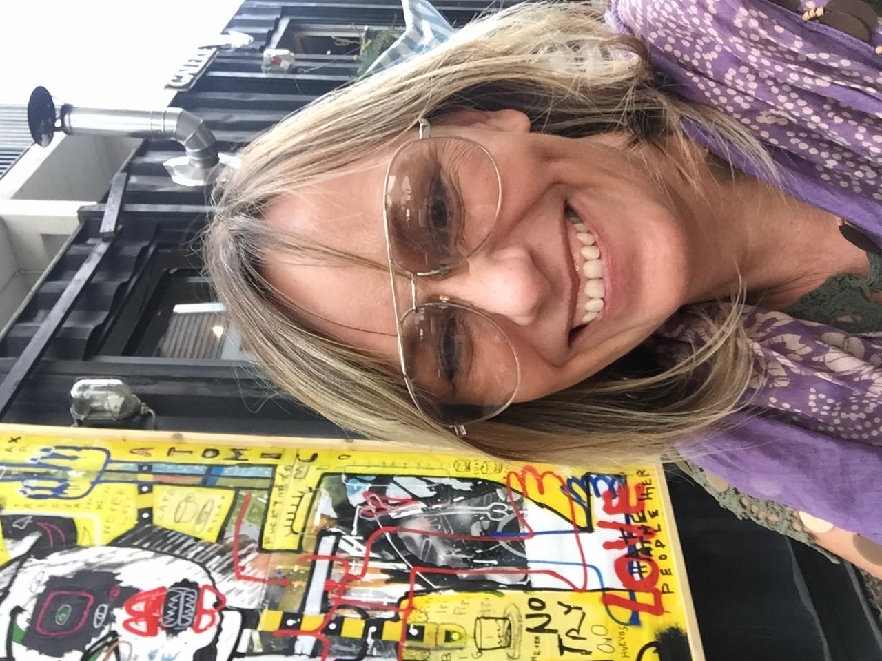 Uśmiechnięta kobieta w okularach pozuje z abstrakcyjnym obrazem na tle czarnego kontenera z metalową rurą, w jasnym, naturalnym świetle.