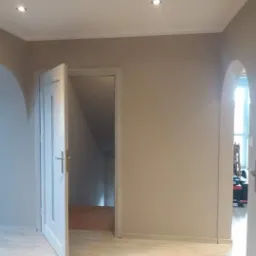 Świeżo pomalowane wnętrze w odcieniach beżu, z otwartymi białymi drzwiami i widocznym wejściem na schody, wykończone listwami przypodłogowymi i dwoma punktowymi lampami sufitowymi, z widocznym...