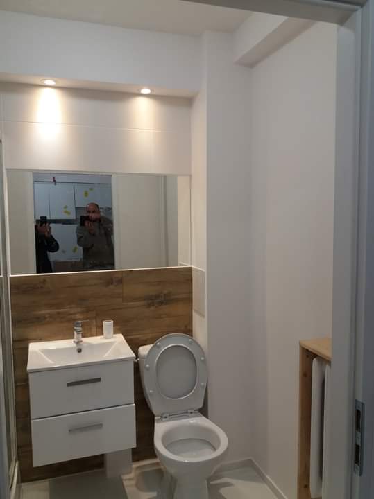 Nowoczesna, jasna łazienka po remoncie z toaletą, białą szafką z umywalką i lustrem, w którym odbija się osoba robiąca zdjęcie. Ściana za umywalką wyłożona płytkami imitującymi drewno.