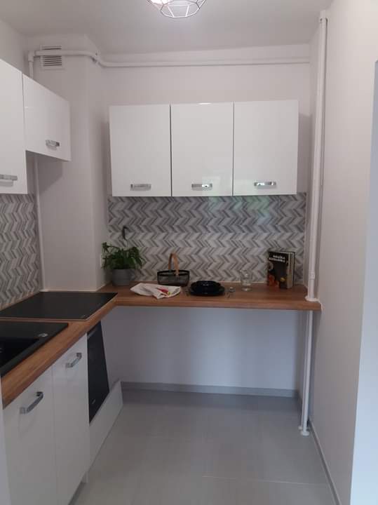 Nowoczesna, jasna kuchnia z białymi, błyszczącymi szafkami, blatem z jasnego drewna i mozaiką w odcieniach szarości na ścianie, uzupełniona o dekoracyjne elementy jak roślina w doniczce i książka...