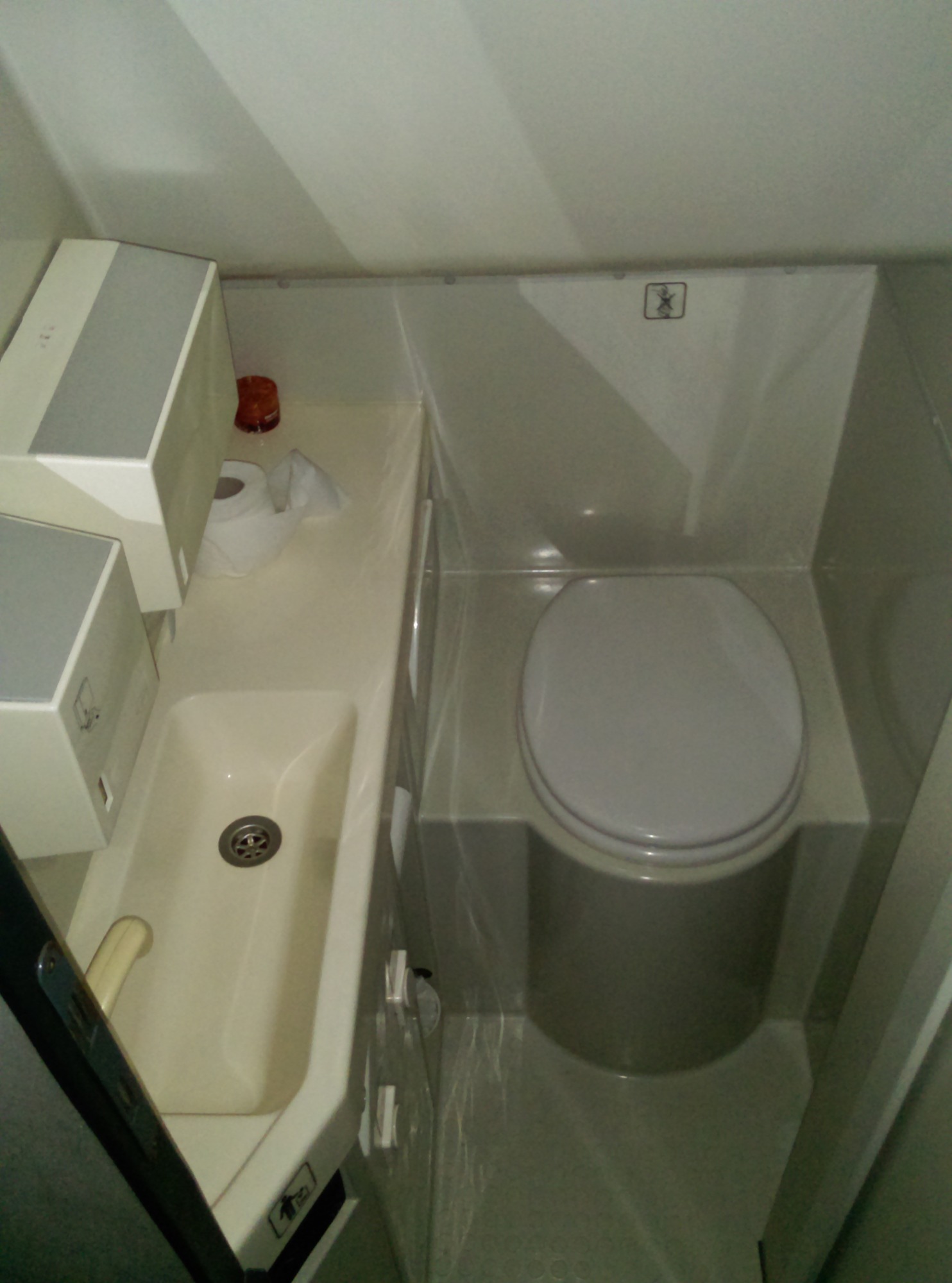 Wnętrze toalety w pojeździe z umywalką, dozownikiem mydła, papierem toaletowym i znakiem informacyjnym.