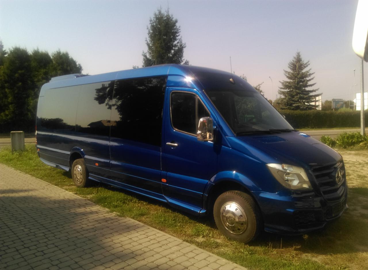 Niebieski, lśniący bus Mercedes Sprinter z przyciemnianymi szybami, zaparkowany na trawie obok chodnika z kostki brukowej w słoneczny dzień.