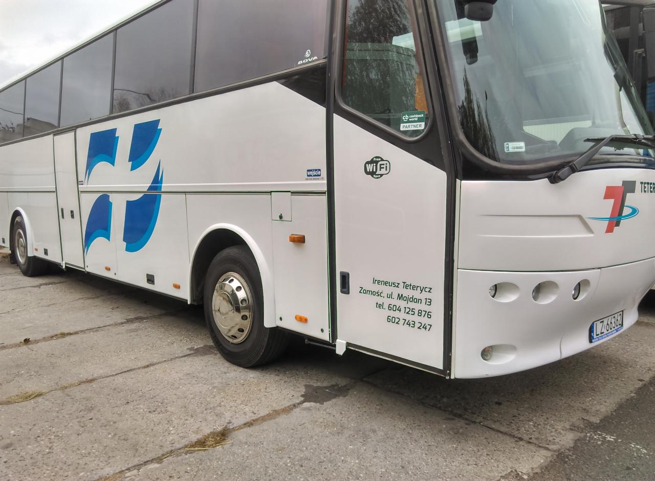 Biały autokar z niebieskim logo firmy na boku, zaparkowany na betonowym placu, widoczne oznaczenie Wi-Fi oraz dane kontaktowe firmy na drzwiach.