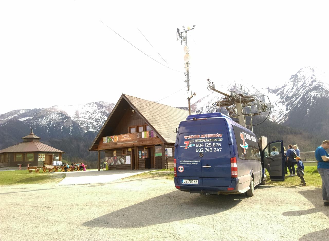 Niebieski bus z otwartymi drzwiami na tle drewnianego budynku, kolejki linowej i górskiego krajobrazu z ośnieżonymi szczytami. Na pojeździe widoczne dane kontaktowe firmy transportowej.