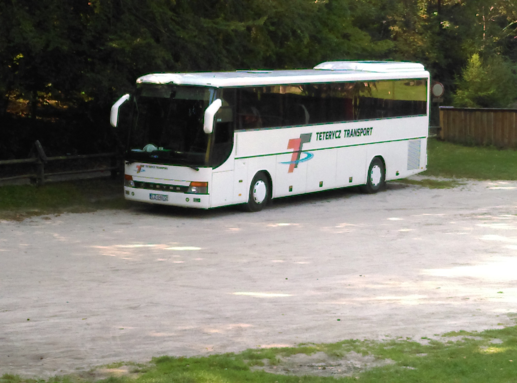 Biały autokar firmy Teterycz Transport zaparkowany na żwirowym placu z zielonymi drzewami w tle.