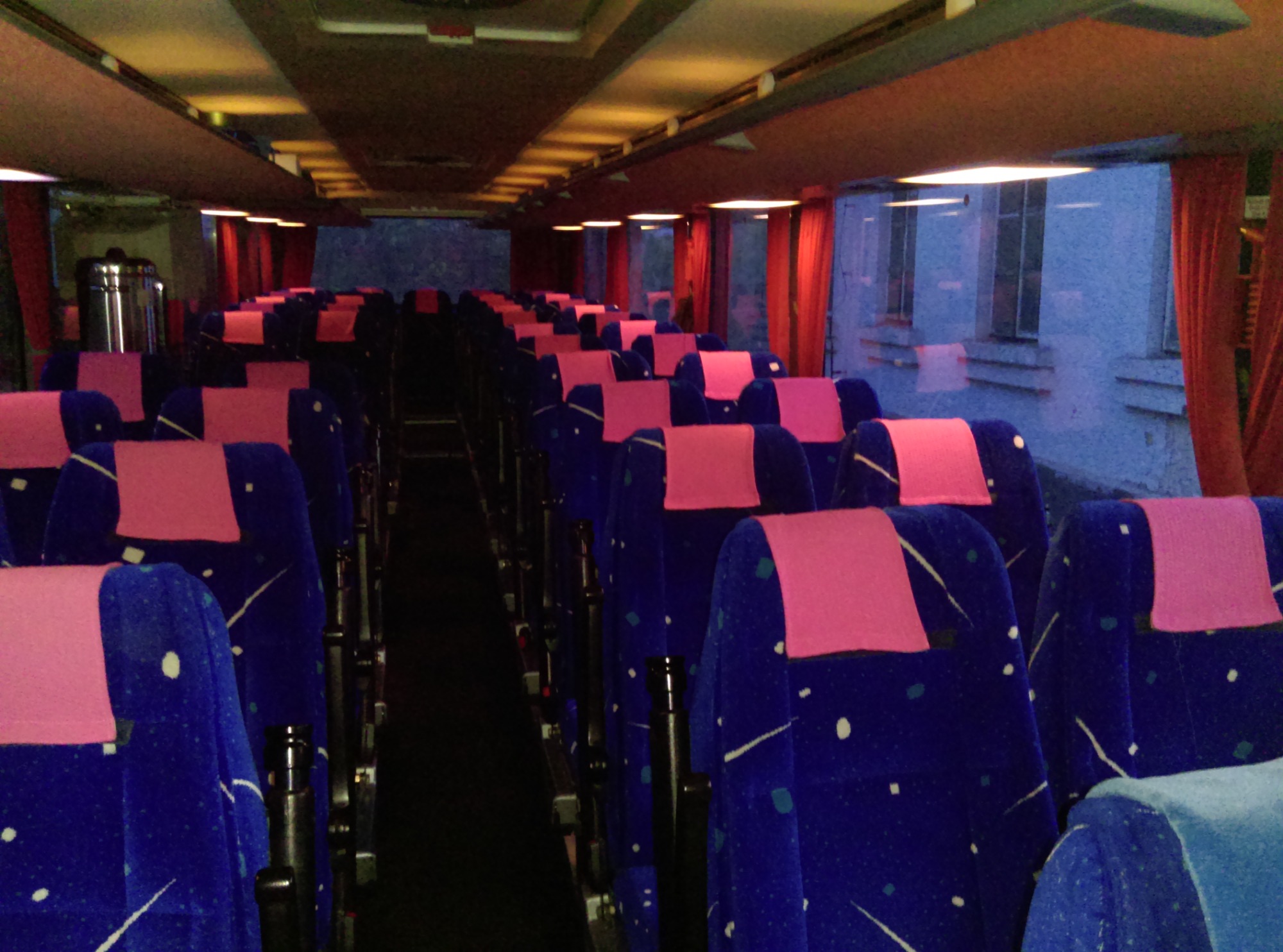 Wnętrze autobusu Setra z rzędami niebieskich foteli z różowymi zagłówkami, widok z perspektywy przejścia, okna z czerwonymi zasłonami w tle.