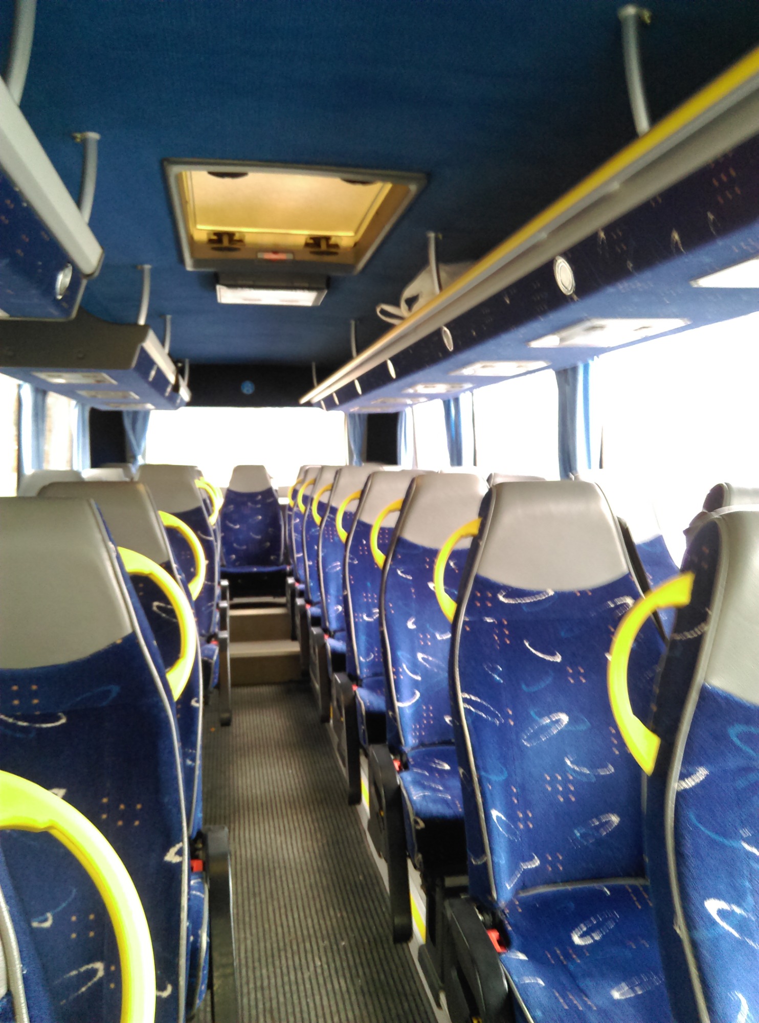 Wnętrze autobusu z rzędami niebieskich siedzeń w abstrakcyjne wzory, żółte uchwyty i szara tapicerka zagłówków, widok w kierunku przedniej części pojazdu.