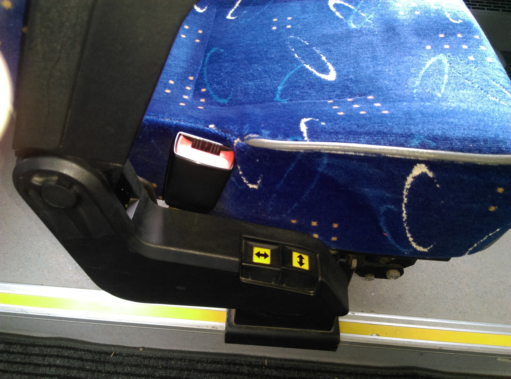 Niebieski fotel z regulacją i pasami bezpieczeństwa w autobusie, widoczne żółte strzałki na panelu regulacji