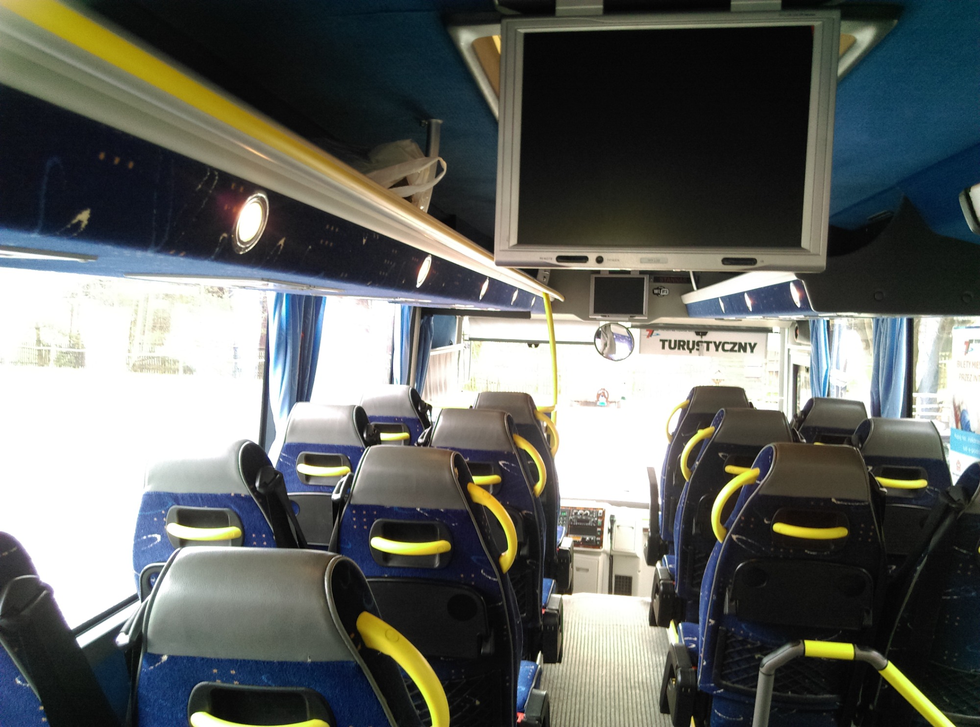 Wnętrze autobusu turystycznego z rzędami niebieskich foteli z żółtymi uchwytami, zamontowany monitor i oświetlenie sufitowe, widok z perspektywy pasażera w kierunku przedniej szyby.