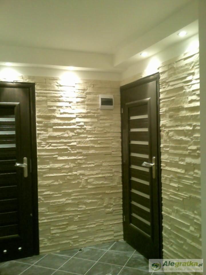Korytarz z dekoracyjną ścianą z imitacji kamienia, oświetleniem punktowym i dwoma ciemnymi drzwiami z poziomymi przeszkleniami.