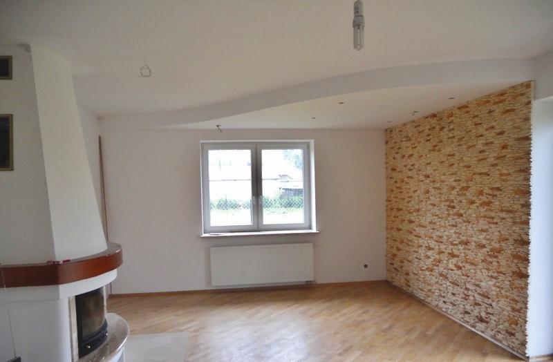 Jasne wnętrze pokoju z drewnianą podłogą, ścianą wyłożoną dekoracyjnym kamieniem, kominkiem z drewnianym blatem i oknem z grzejnikiem.