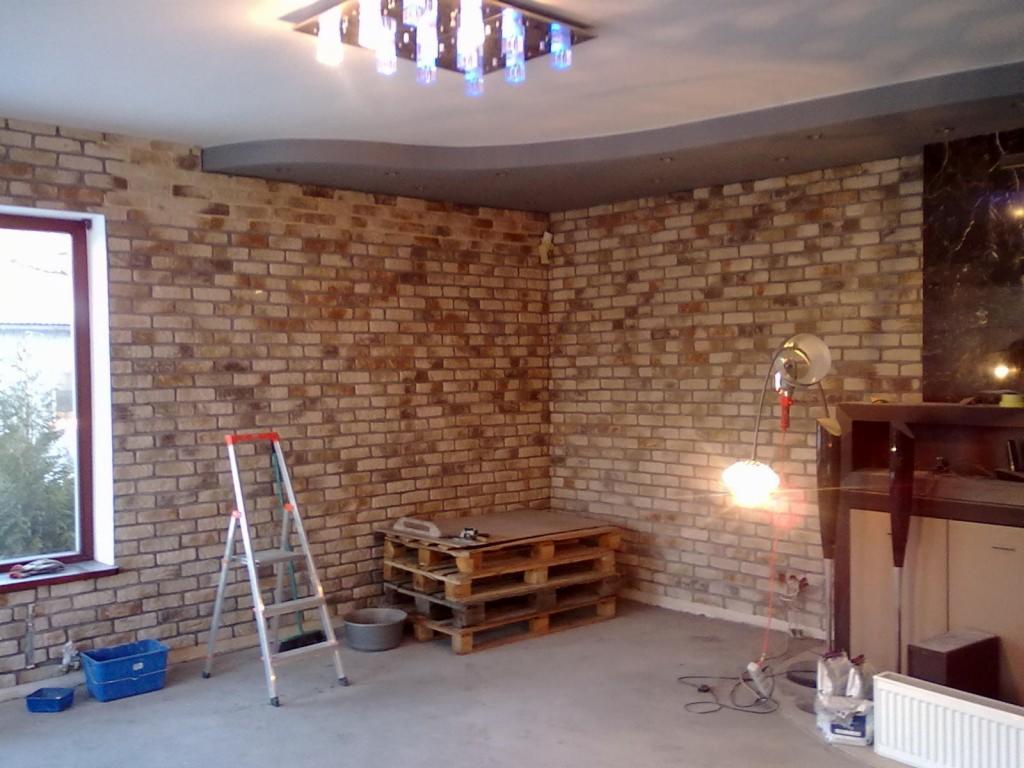 Ściana wyłożona cegłą dekoracyjną w trakcie prac wykończeniowych, widoczna drabina, palety, lampa halogenowa i grzejnik.