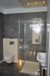 Nowoczesna łazienka z kabiną prysznicową typu walk-in, podwieszaną toaletą i ciemnymi płytkami na ścianach oraz podłodze. Oświetlenie punktowe w suficie i dekoracyjne świeczki na podłodze. Widoczny...