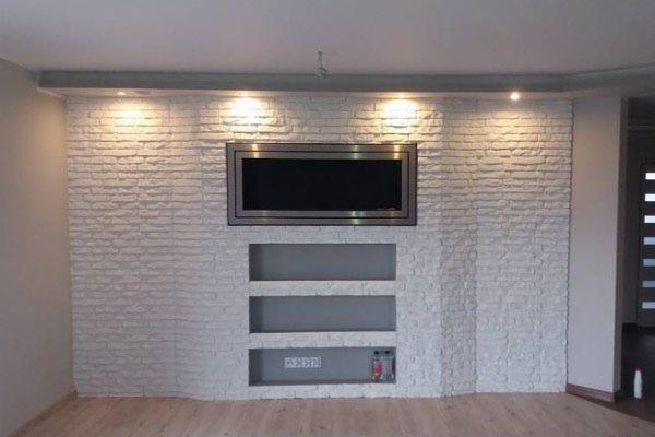 Ściana z białej cegły dekoracyjnej z wnękami, podświetlona punktowo, z wbudowanym telewizorem w srebrnej ramie.