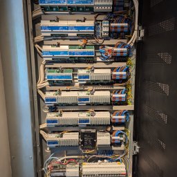 Smart Instal - Otwarta szafa sterownicza z precyzyjnie ułożonymi modułami automatyki budynkowej Lutron HomeWorks DALI i QS, kolorowymi przewodami oraz zabezpieczeniami. Widoczne szczegóły instalacji elektrycznej.