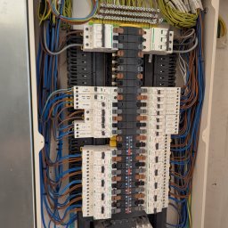 Smart Instal - Otwarta skrzynka elektryczna z rzędami wyłączników Schneider, licznymi kolorowymi przewodami (niebieskie, brązowe, żółto-zielone) i szynami zbiorczymi, z widocznymi etykietami obwodów.