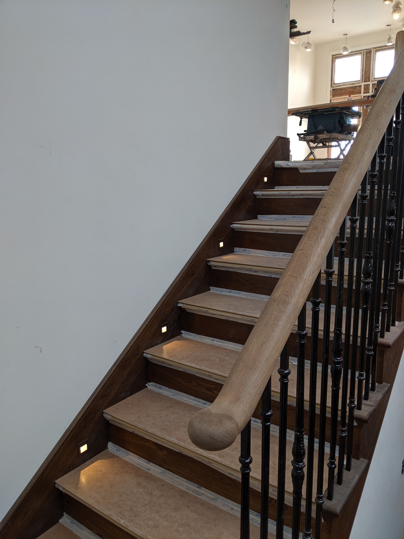 Schody drewniane z czarnymi, ozdobnymi balustradami i wbudowanym oświetleniem LED na każdym stopniu, widoczne zabezpieczenia folią malarską, w tle pomieszczenie z narzędziami.