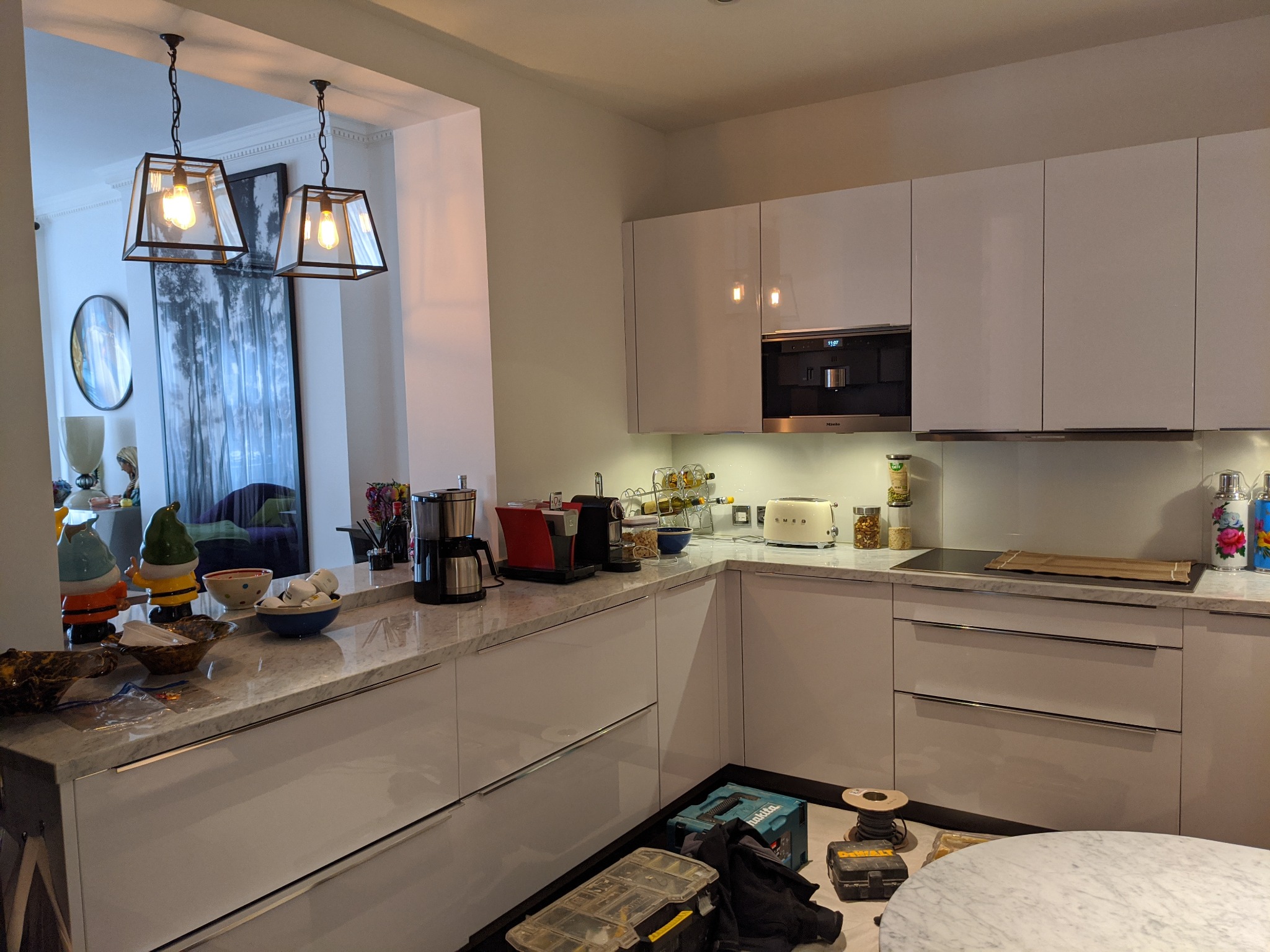 Nowoczesna kuchnia z białymi szafkami i marmurowym blatem, z widocznymi narzędziami elektrycznymi na podłodze, sugerującymi trwający montaż oświetlenia.