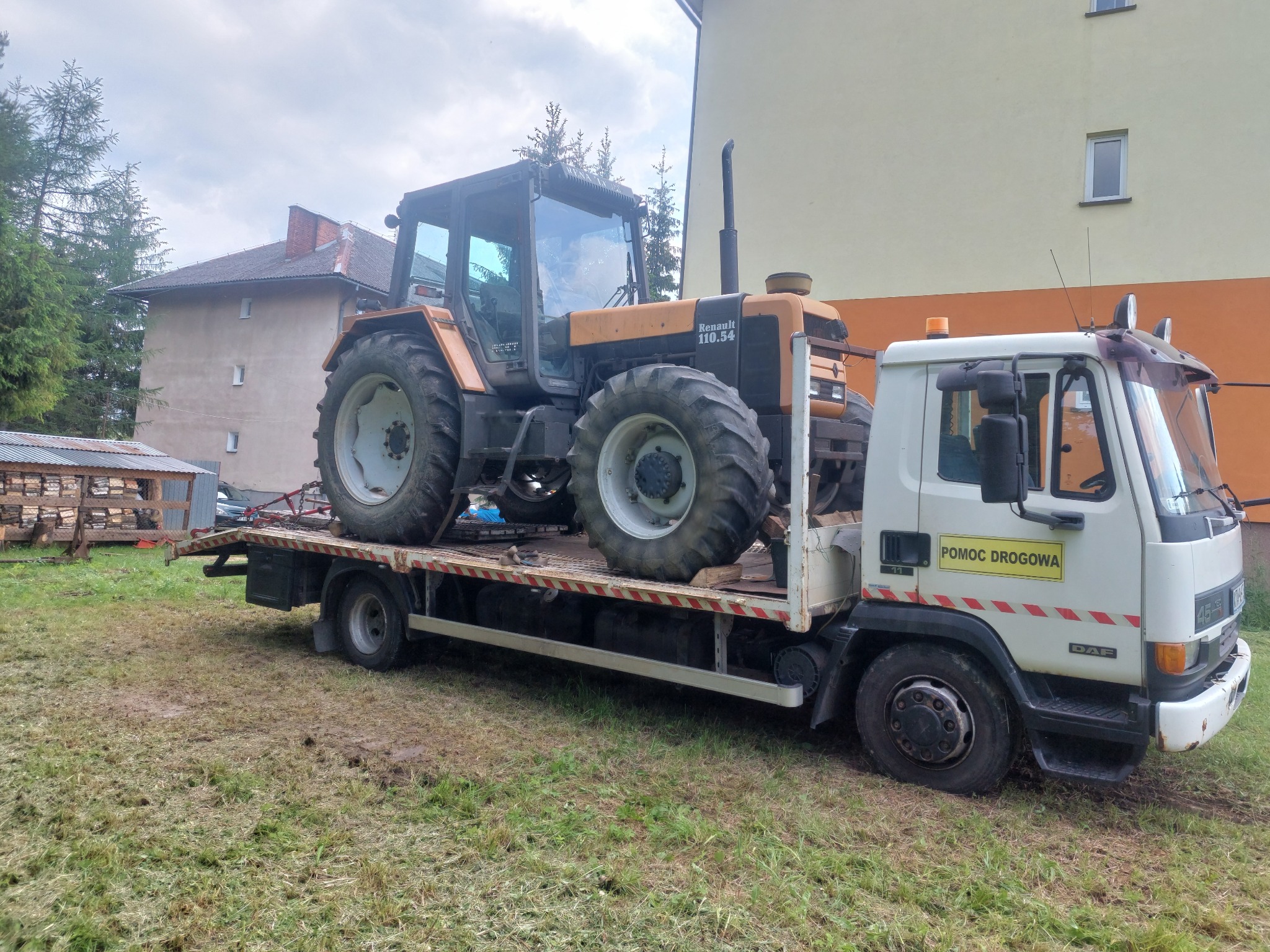 Laweta DAF z napisem 'Pomoc Drogowa' przewozi ciągnik rolniczy Renault 110.54 na tle budynków.