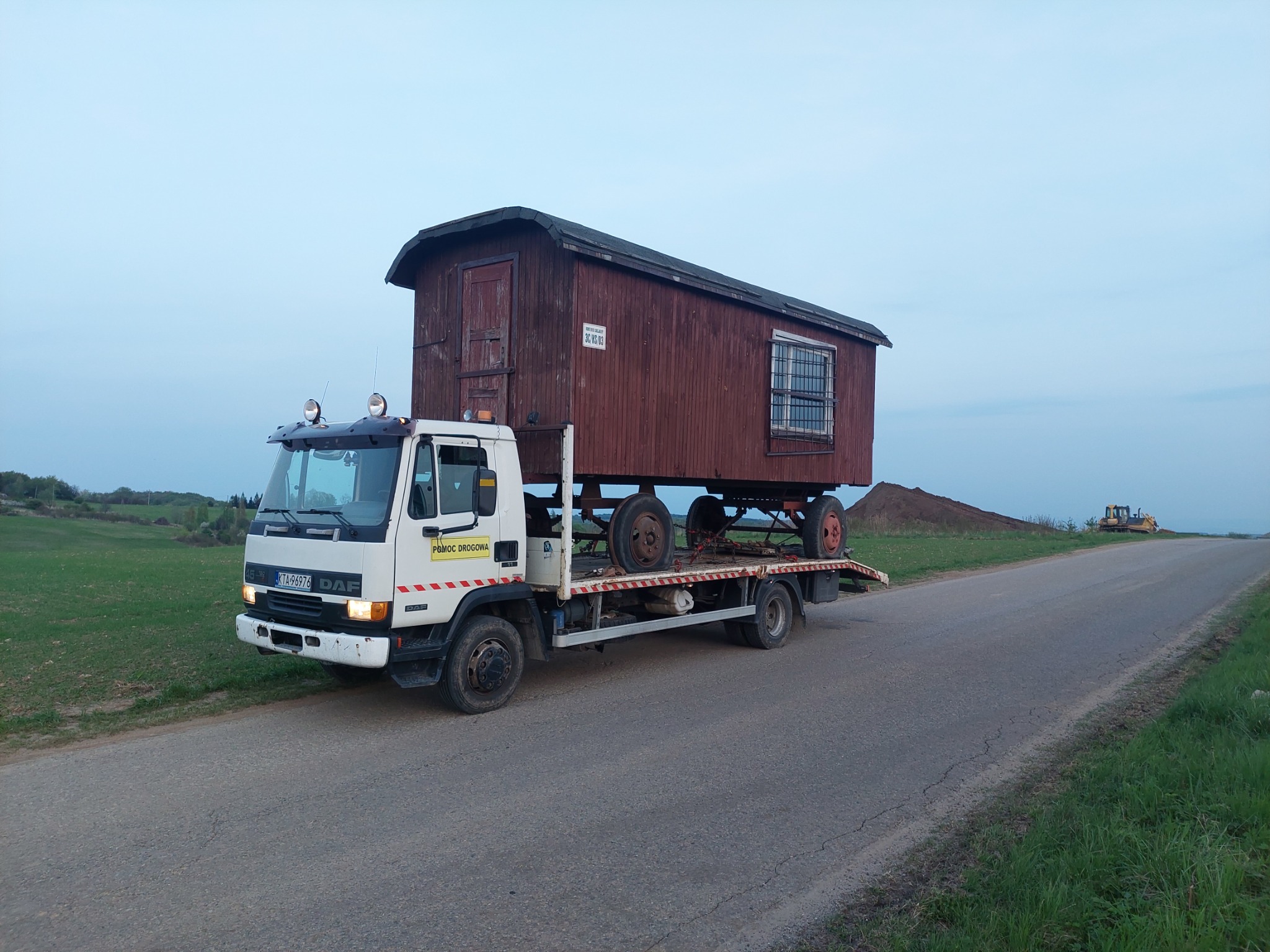 Biała laweta DAF z napisem 'Pomoc Drogowa' przewozi na platformie drewniany barak z oknem w kratę, ustawiony na kołach. W tle widoczny krajobraz z polem i koparką.