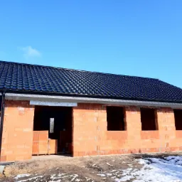 Budynek w stanie surowym otwartym z czarną dachówką, widoczne bloczki z betonu komórkowego oraz resztki śniegu na ziemi.