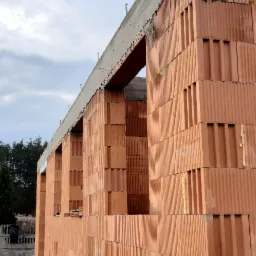 Surowy stan domu z czerwonej cegły ceramicznej z widocznymi otworami okiennymi i betonowym wieńcem, w tle drzewa i pochmurne niebo.