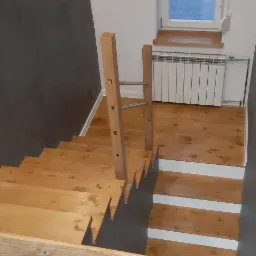 Drewniane schody z jasną balustradą na tle szarej ściany, widok z góry na stopnie z białymi podstopnicami, w tle grzejnik i okno.