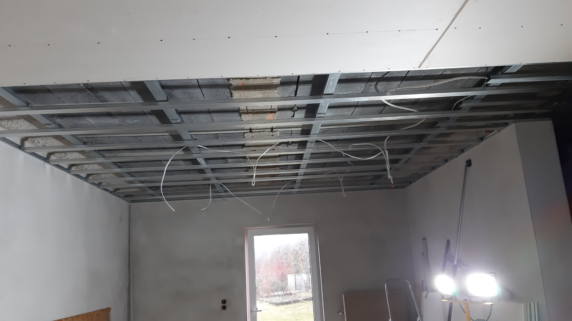 Wnętrze w trakcie budowy: sufit z widoczną konstrukcją metalową, częściowo zakryty płytami kartonowo-gipsowymi, wystające przewody elektryczne, widok na okno i oświetlenie budowlane.