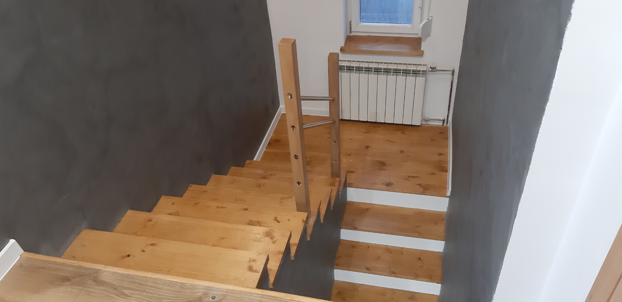 Drewniane schody z jasną balustradą na tle szarej ściany, widok z góry na stopnie z białymi podstopnicami, w tle grzejnik i okno.