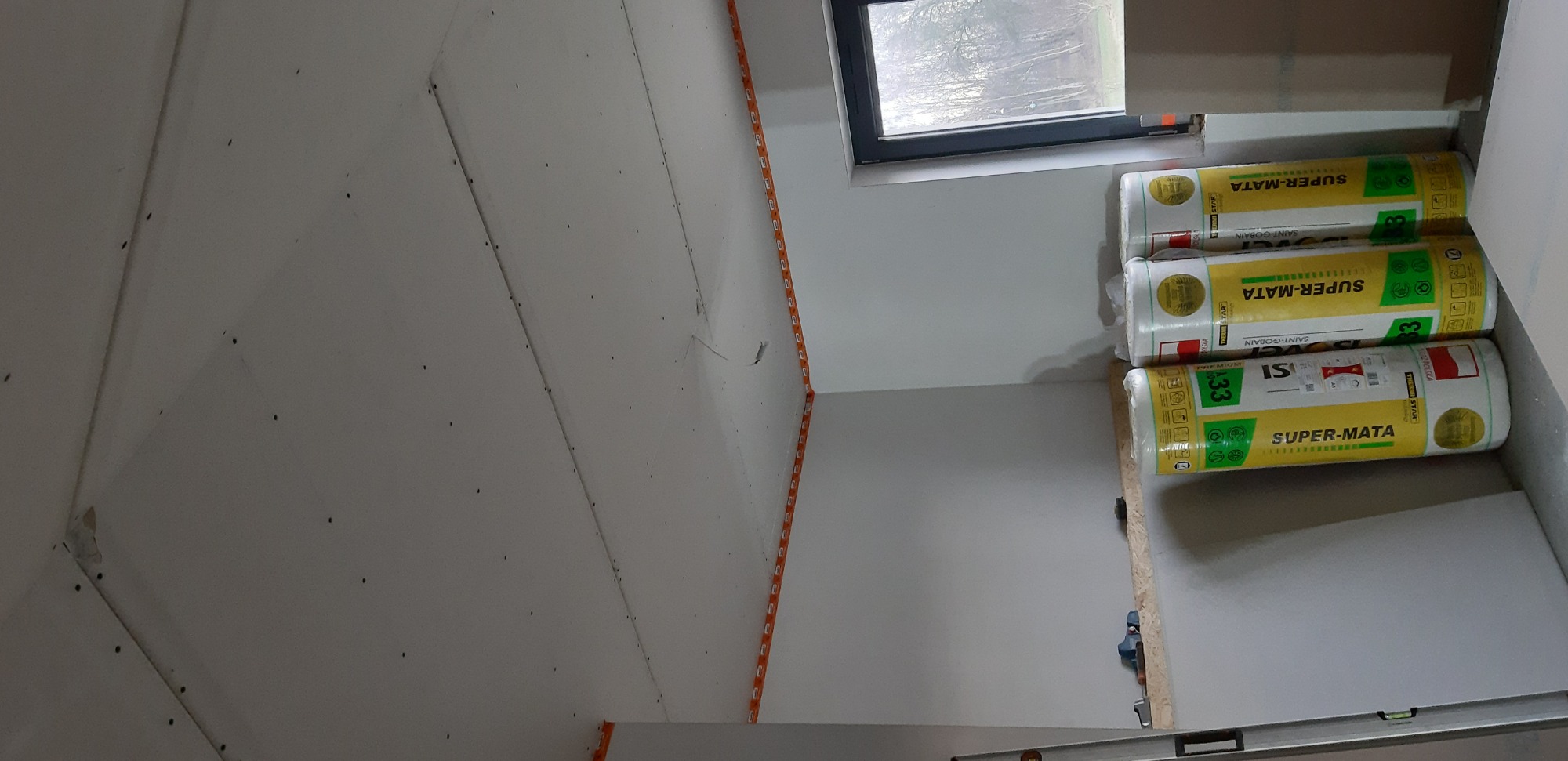 Wnętrze w trakcie ocieplania: widoczne płyty kartonowo-gipsowe na ścianach, profile montażowe z pomarańczowymi znacznikami oraz rolki wełny mineralnej Super-Mata Saint-Gobain w folii, przygotowane...