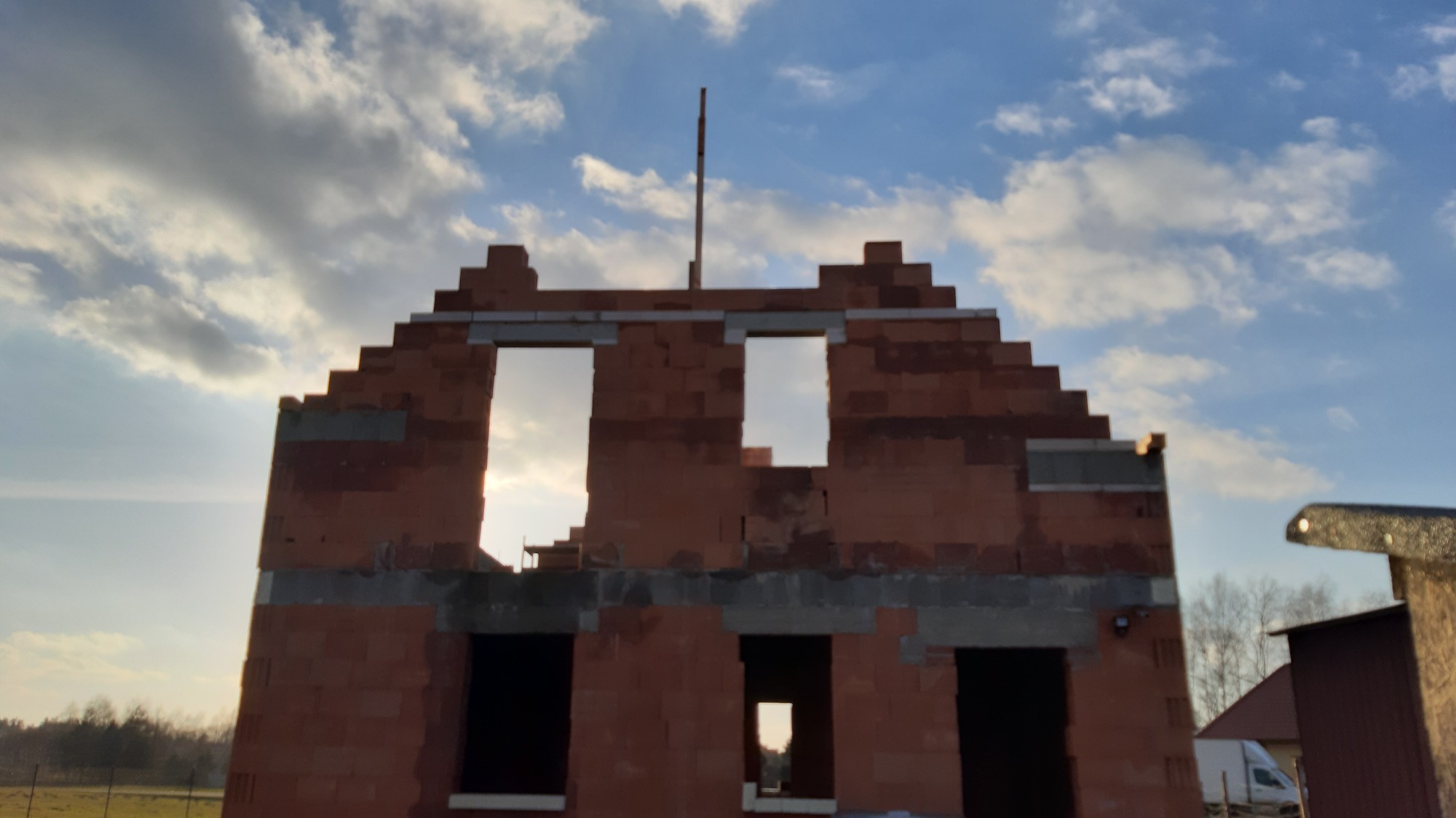 Surowy stan domu murowanego z czerwonej cegły, widoczne otwory okienne i drzwiowe, zbrojenie na wysokości stropu, w tle błękitne niebo z chmurami.