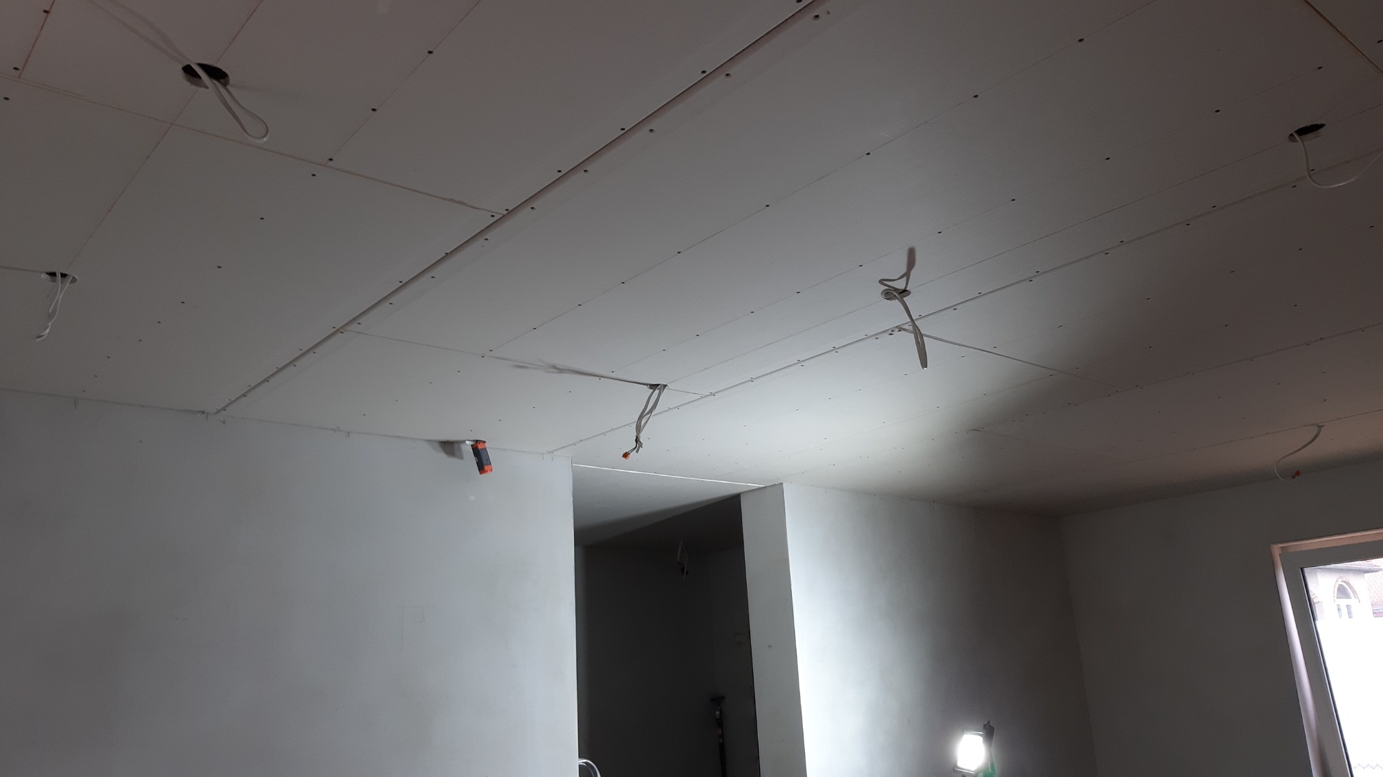 Wnętrze w trakcie wykańczania: sufit z płyt gipsowo-kartonowych z widocznymi przewodami elektrycznymi i otworami na oświetlenie, ściany w stanie surowym, widok na przejście do kolejnego pomieszczenia.