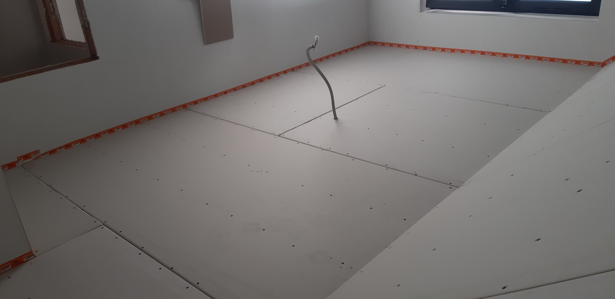 Wnętrze poddasza z zamontowanymi płytami gipsowo-kartonowymi, widoczne kable instalacji elektrycznej wystające z podłogi, pomarańczowa taśma uszczelniająca na obrzeżach, okno w tle.