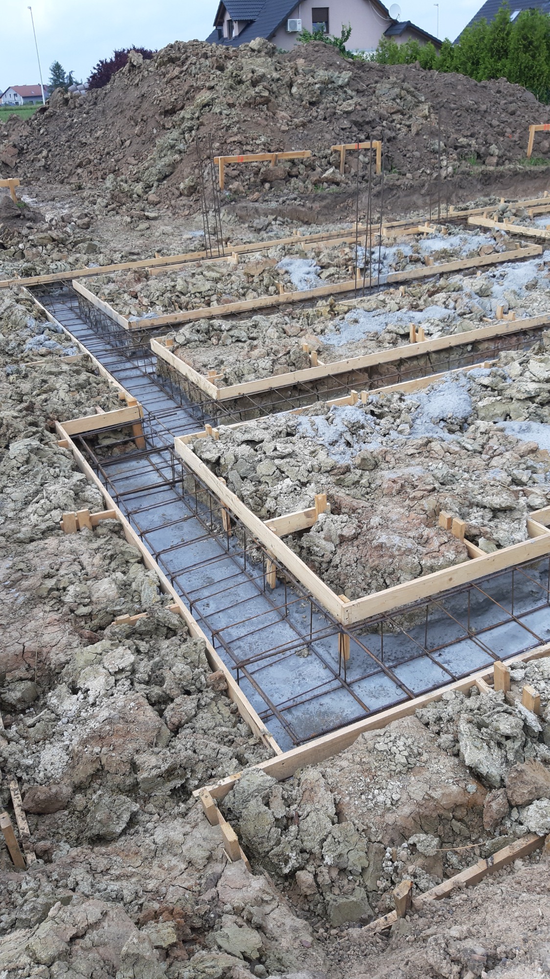 Zalane betonem szalunki zbrojeniowe fundamentów domu jednorodzinnego, widok z góry na plac budowy z wykopem i stertą ziemi w tle.