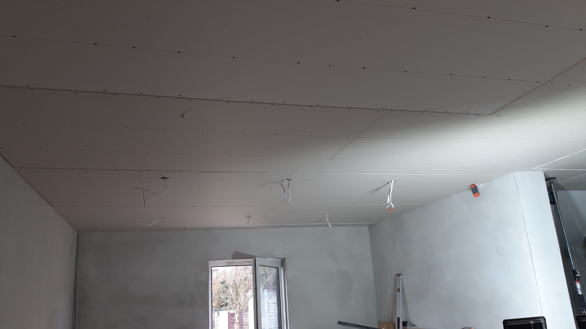 Wnętrze w stanie surowym: zamontowane płyty gipsowo-kartonowe na suficie, wystające kable instalacji elektrycznej, widoczne narzędzia i poziomica przy ścianie, otwarte okno z widokiem na drzewa.