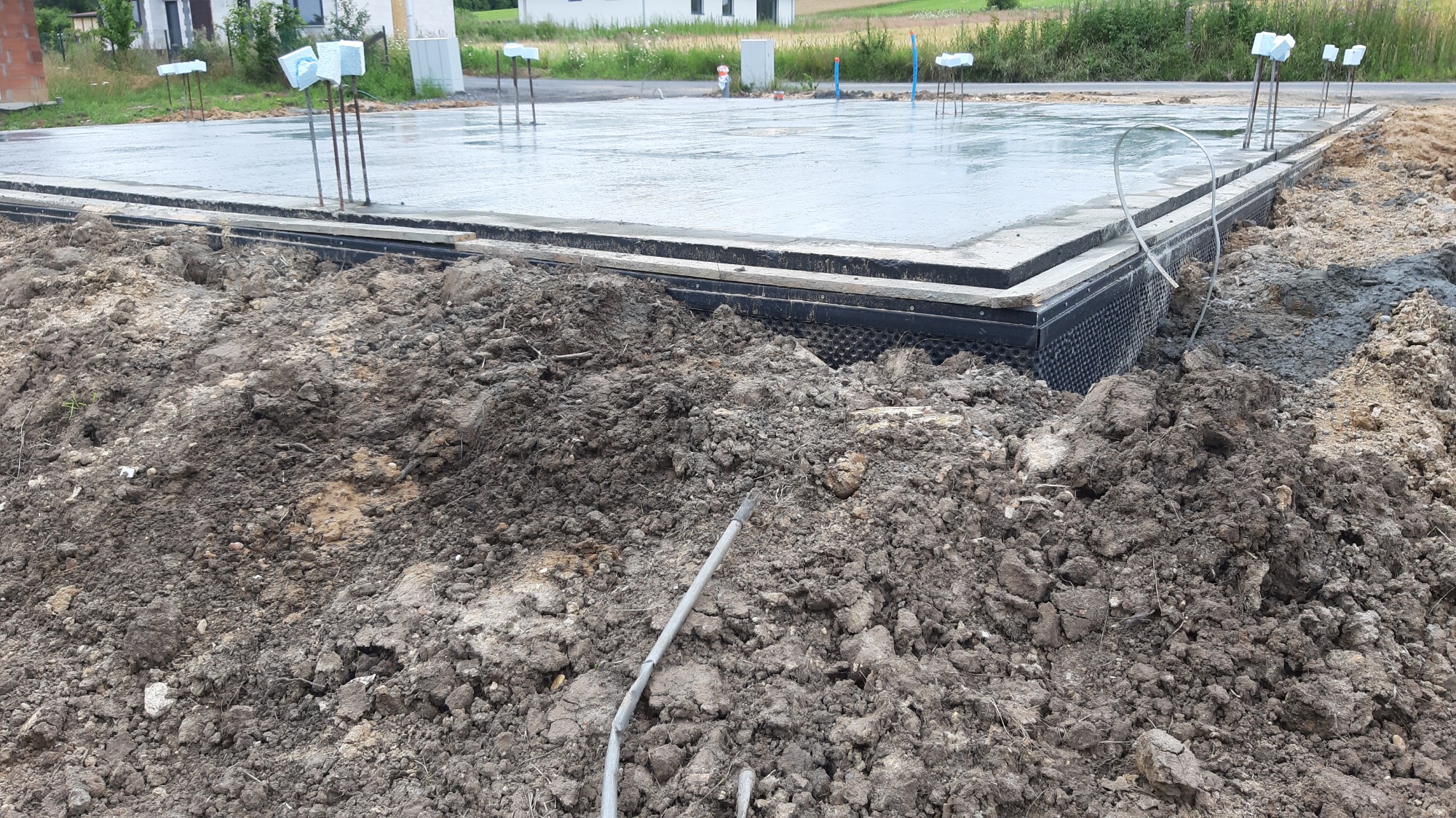 Wylany fundament z hydroizolacją, widoczne wystające pręty zbrojeniowe i kopiec ziemi w pierwszym planie.